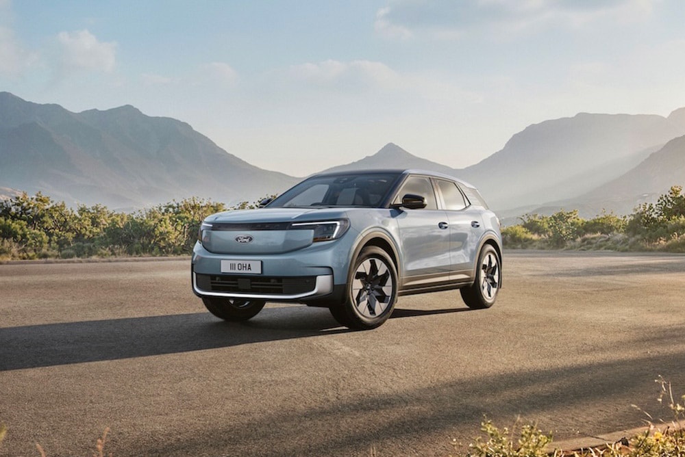 Front view of a light blue Ford Explorer EV parked on a paved road with mountains and trees.