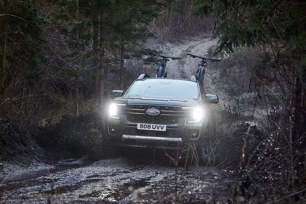 A grey Ford Ranger driving up a muddy hill in a forest carrying 2 bikes in the bed, seen from a front profile angle.