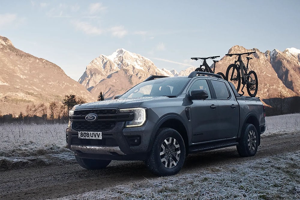 A grey Ford Ranger driving through a frosty field surrounded by mountains carrying 2 bikes in the bed, seen from a front three-quarter angle.