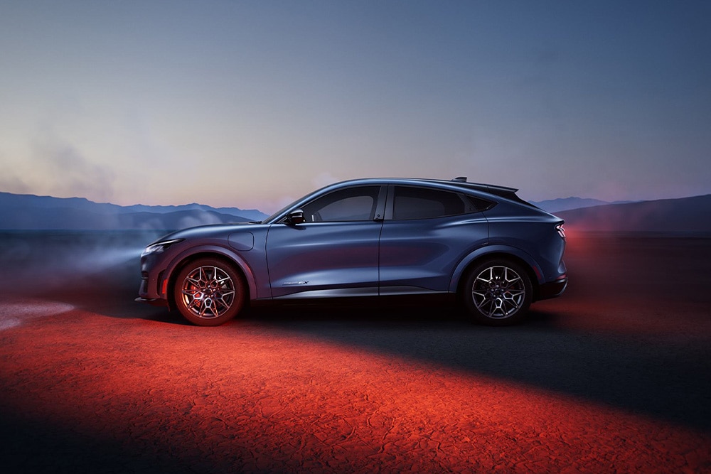 A side profile of a blue Ford Mustang Mach-E parked in a desert landscape at twilight, with red taillights illuminating the ground.