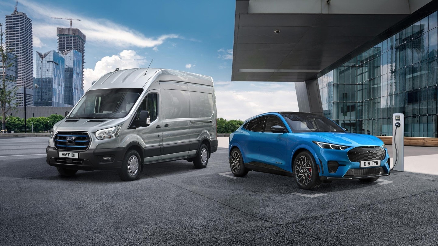 A grey Transit and blue Mach-E charging in front of a building