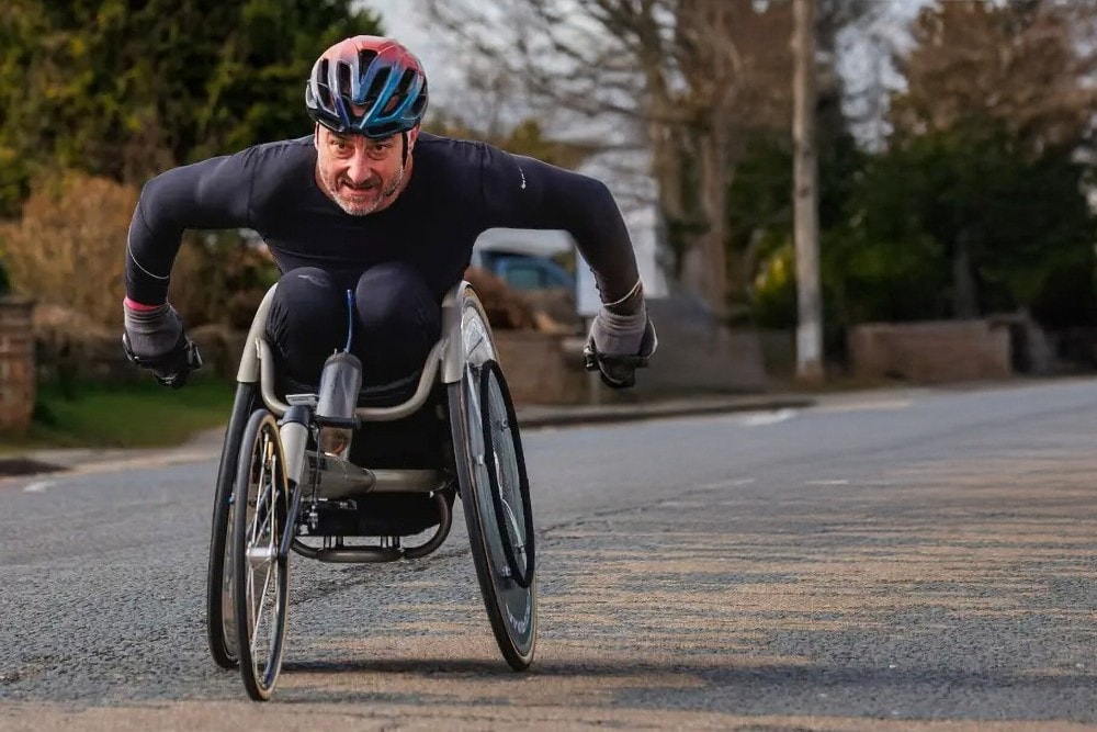 Jason Richards in a racing wheelchair wearing a helmet, propelling themselves along a road